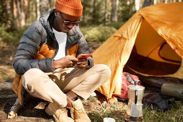 Picture of african man in stylish warm clothes and sunglasses using smartphone sitting in wild forest near orange tent in campsite, camping in wild nature surfing mobile internet as freelance worker