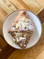Fresh almond croissant. Pastry. White ceramic plate with freshly baked almond croissant on a wooden background. Directly above. having breakfast in a cafe.