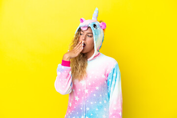 Girl with curly hair wearing a unicorn pajama isolated on yellow background yawning and covering wide open mouth with hand