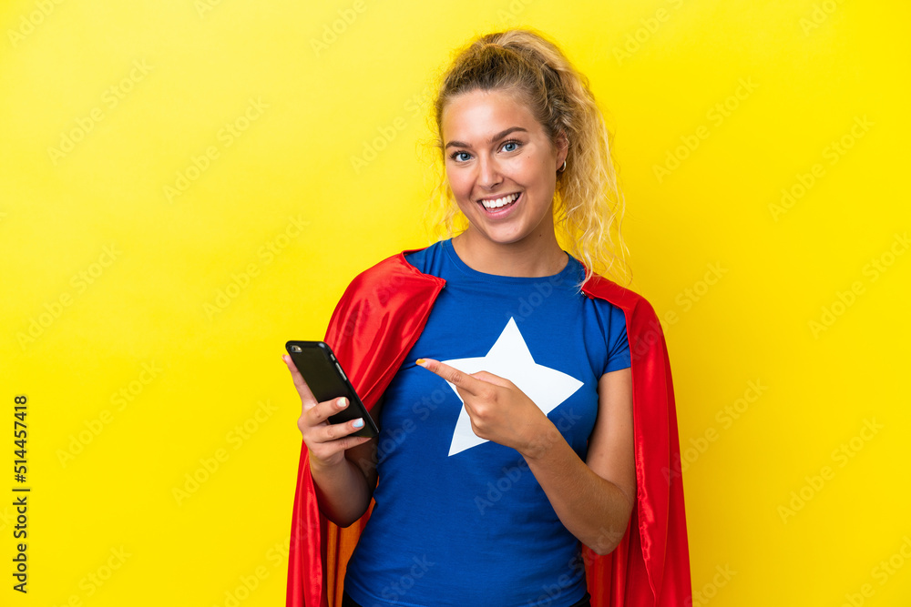 Wall mural Super Hero woman isolated on yellow background using mobile phone and pointing it