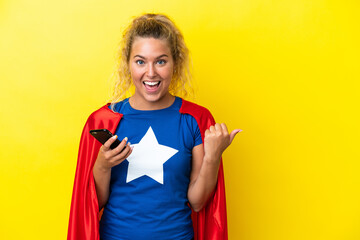 Super Hero woman isolated on yellow background using mobile phone and pointing to the lateral