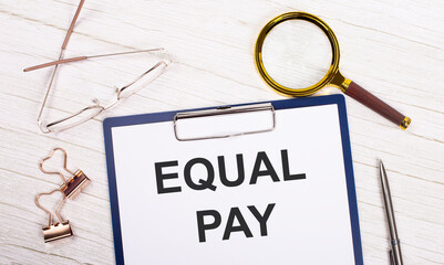 A tablet with the text EQUAL PAY on white paper, a magnifier, glasses, golden paper clips and a pen lie on a white office table. Top view, flat lay.