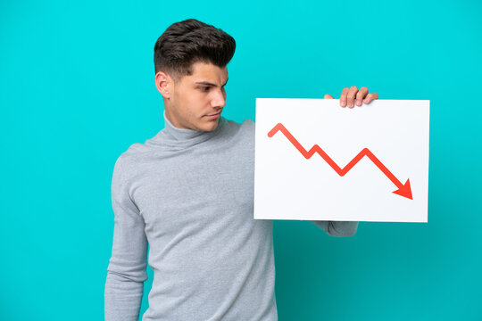 Young handsome caucasian man isolated on blue bakcground holding a sign with a decreasing statistics arrow symbol with sad expression