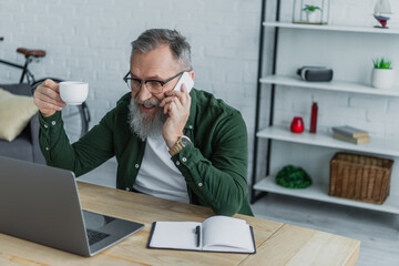 bearded senior man in eyeglasses holding cup and talking on smartphone near laptop.