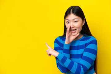 Young Chinese woman isolated on yellow background pointing to the side to present a product and whispering something