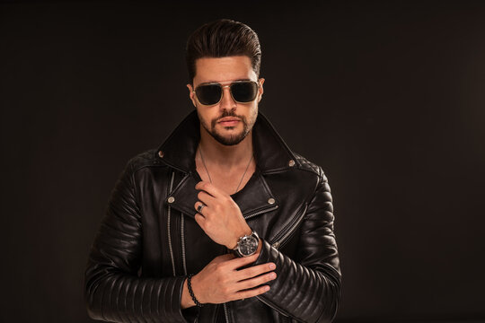 Fashion photo of handsome man wearing black leather jacket, sunglasses, hand watch, posing on dark studio background.