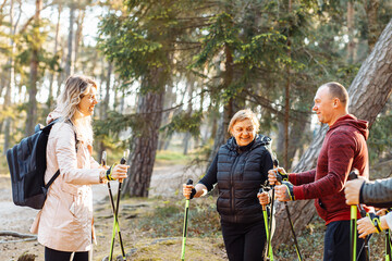 Smiling woman sports trainer with group of people talking, explaining posture for Nordic walking with sticks in forest