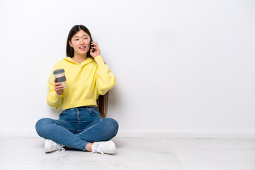Young Chinese woman sitting on the floor isolated on white wall holding coffee to take away and a mobile