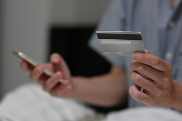 Close up of Asian male with nighdress  holding a back gray credit card and a smartphone for online shopping. Cashless and e-commrece concepts.