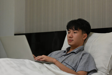 Portrait of Asian workaholic male using his computer laptop on bed for work before he sleeps. Business and financial concepts.