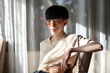 Portrait of LGBT Asian slim male with crop top cream shirt and orange glasses looking at camera and...