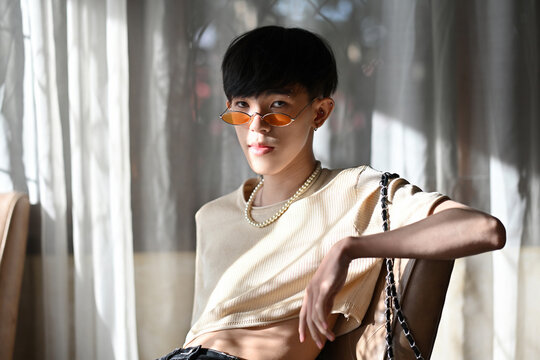 Portrait of LGBT Asian slim male with crop top cream shirt and orange glasses looking at camera and sitting on chair with a pose and sunlight. People lifestyle fashion LGBTQ concept.
