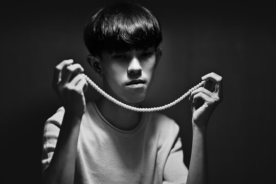 Black and white photo of Asian male with black short hair holding and looking at his necklace with sad eyes. Abstract high exposure contrast shadow.