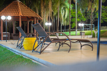 Lounger chair next to a swimming pool, palm trees, lights, hut, grass, green, night, tiles.