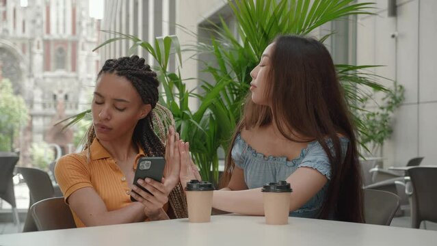 Head shot smiling young African American girl sitting on comfortable chear, obsessed with smartphone, using mobile apps, playing mobile games or shopping in internet store ignoring Asian girlfriend