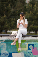 Naklejka premium A beautiful blonde with long hair in a white outfit sits on a bench and flips through the phone