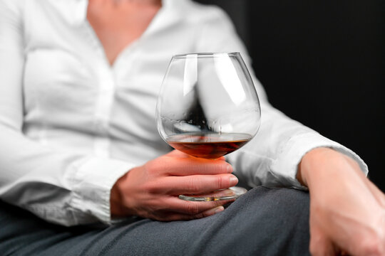 Drinking glass filled with alcohol held by woman in business clothes sitting with her legs crossed. Woman's image blurred. Concept for depression, sadness, alcoholism, drinking problems.