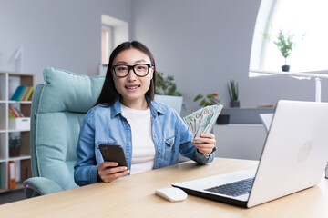 Fototapeta na wymiar Portrait of young beautiful freelancer woman looking at camera and smiling holding phone and american money dollars, online win