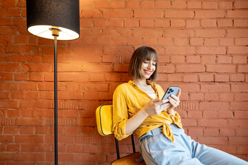 Young stylish woman using smart phone while sitting on chair near floor lamp on brick wall...