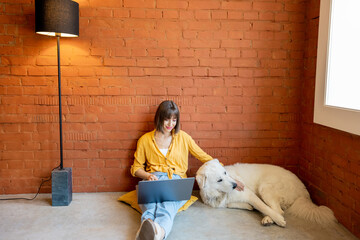 Young woman working on laptop computer while sitting with her cute dog on brick wall background at home. Work from home and friendship with pets concept