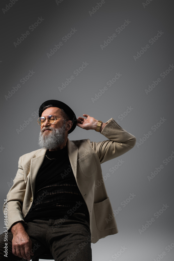 Poster bearded senior man in eyeglasses and jacket adjusting derby hat on grey.