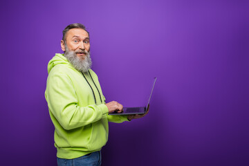 smiling senior man with beard and grey hair using laptop on purple background.