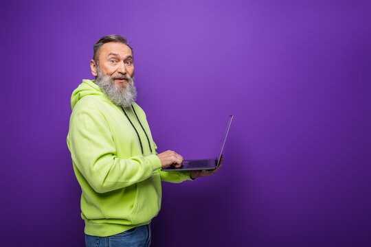 Smiling Senior Man With Beard And Grey Hair Using Laptop On Purple Background.