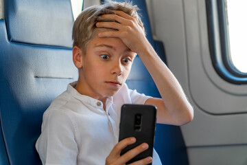 Amazed caucasian teenage holds his forehead with his hand and looks in surprise at phone screen while riding commuter train