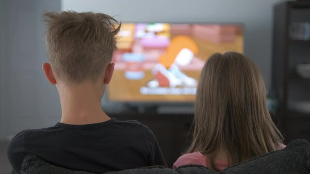 Rear view of brother and sister watching TV at home. Children sitting on sofa enjoying cartoons together