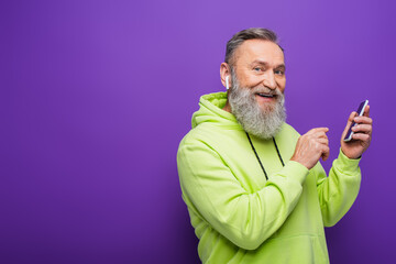 happy senior man in green hoodie listening music in wireless earphone and holding smartphone on purple.