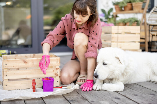 Young woman painting wooden box in pink color, doing housework while sitting with her dog on terrace of her house. DIY concept. Idea of friendship with pets
