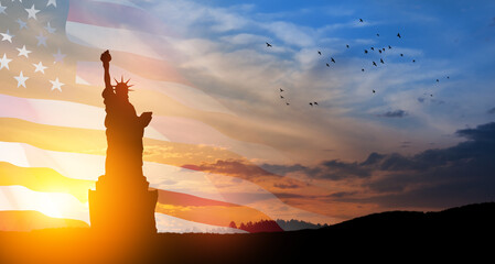 Statue of Liberty with a large american flag and sunset sky with flying birds on background.