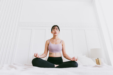 Fototapeta premium Sporty Asian woman practicing yoga on a white bed, doing Ardha Padmasana exercise, meditating in Half Lotus pose, indoor working out at home, wearing sportswear.