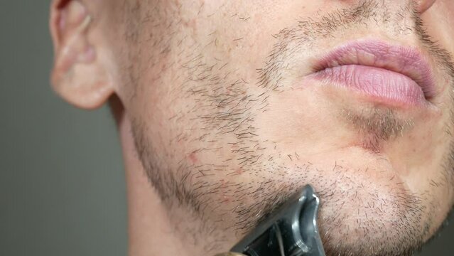 Man is cutting beard by Electric metal razor or dry shaver. Trimmer for cutting. Short, sparse beard on mans face. Hair growth problems. Unshaven bristles on the beard.
