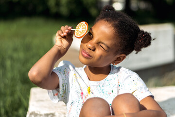 A cute African-American girl covers one eye with a bright fruit lollipop in the park on a summer...