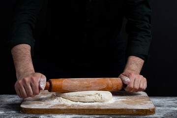Male hands with wooden rolling pin rolls out dough. Close Up view of baker's male hands making...