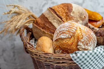 Homemade freshly baked bread, buns and a long loaf in a large wicker basket. Wood rustic table, board, ears, wheat, rye, grain harvest. Rural food cooking