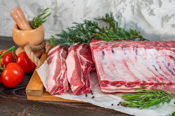 Raw pork loin, bacon with ribs on a wooden board, spice mortar, and pestle, rosemary, tomatoes. Pieces of meat, ingredients for cooking. Protein nutrition, a diet with fresh herbs and vegetables