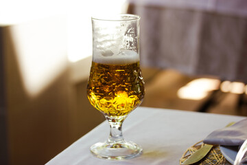 A beer in Tulip glass. Beautiful crystal glass with golden freshly brewed cold wheat beer stands on white tablecloth on table on cafe, restaurant terrace. A refreshing alcoholic beverage in summer day