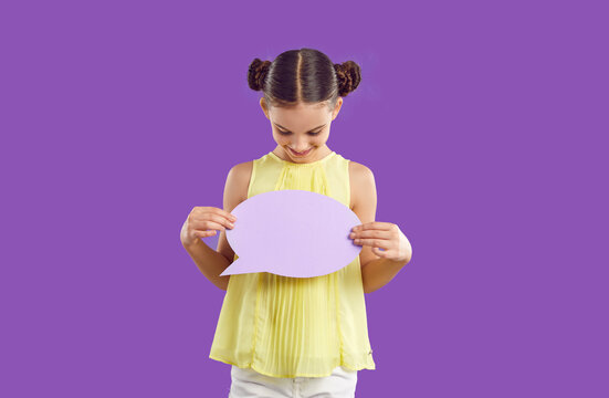Happy smiling elementary school girl with text bubble standing on purple studio background. Cute pretten girl with two bundles on her head looks at empty speech bubble in her hands. Copy space.