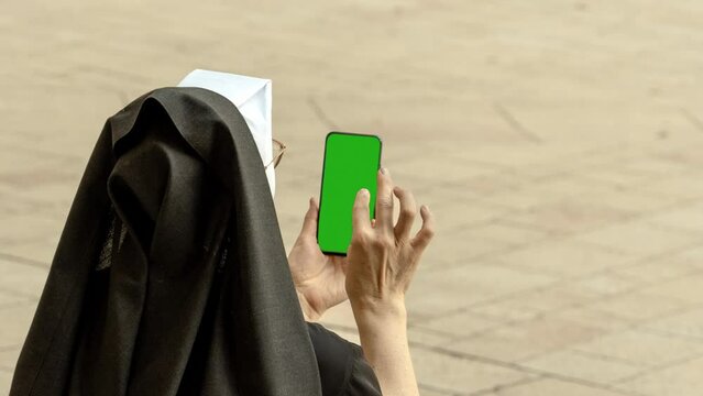 An elderly nun holds a mobile phone with a vertical green screen on a gray square, green screen, smartphone, a mobile phone touch screen display in a female hand. Blank screen for content integration