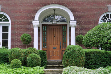 Fototapeta na wymiar Portico entrance of traditional brick house surrounded by shrubbery, with elegant wood grain front door with sidelights