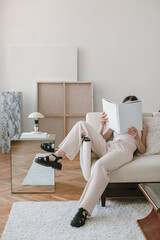 Unrecognizable girl holding a book with a blank cover sitting on a sofa in a stylish apartment. Modern creative interior. Mock up.