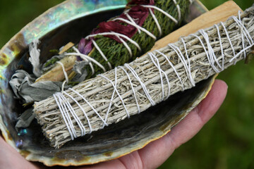 A close up image of a hand holding an abalone shell filled with handmade smudge bundles. 