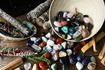 An image of a bowl of healing crystals spilled on a wooden table with healing smudge bundles. 