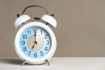 Seven o'clock on the alarm. A white alarm clock is on a white table. The clock hand points to 7...