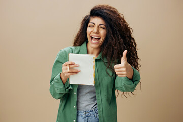 COOL CONCEPT. Overjoyed tanned curly Latin female in casual things with notebook gesture thumb up posing isolated over pastel beige background look at camera. Copy space Mockup Banner. Good offer