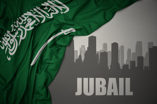 abstract silhouette of the city with text Jubail near waving national flag of saudi arabia on a gray background.
