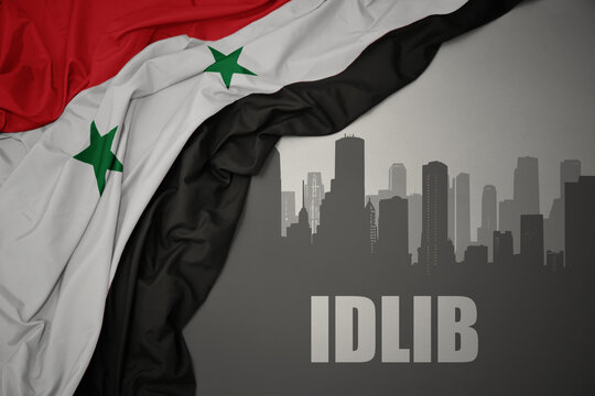 abstract silhouette of the city with text Idlib near waving national flag of syria on a gray background.