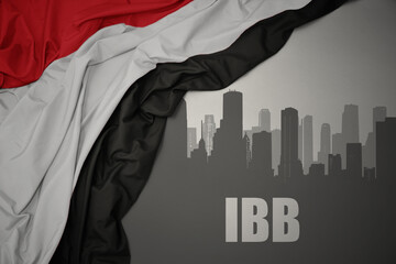 abstract silhouette of the city with text Ibb near waving national flag of yemen on a gray background.